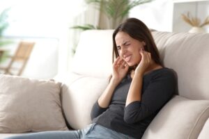 Woman on couch squinting in pain touching hands to jaw